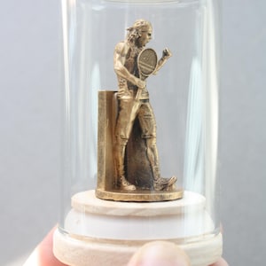 May include: A bronze tennis player figurine inside a glass cloche. The figurine is of a man in tennis attire, holding a racket and celebrating. The cloche sits on a wooden base, and the entire piece is held by a hand.