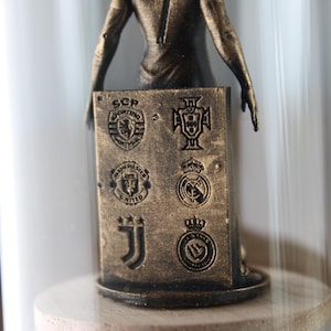May include: A bronze-colored figurine of a soccer player, with the name "RONALDO" and the number "7" on the back. The figurine holds a rectangular plaque with team logos. The figurine is displayed inside a glass dome.
