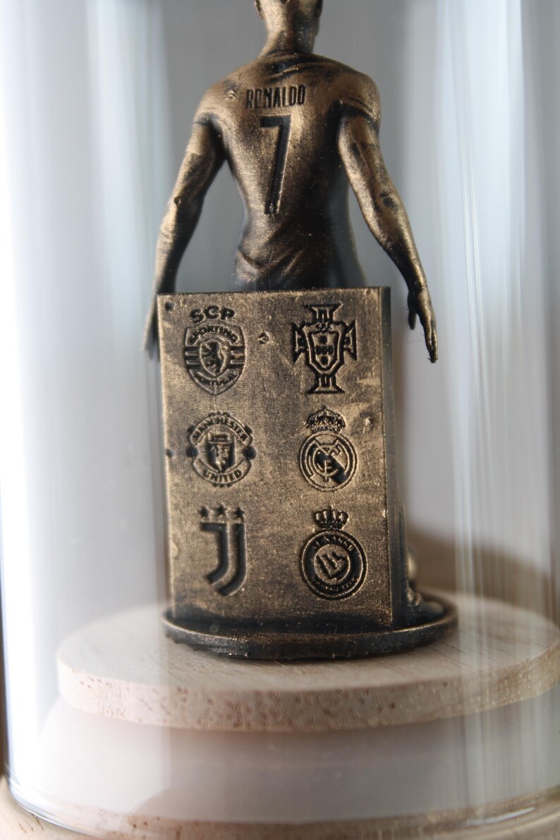 May include: A bronze-colored figurine of a soccer player, with the name "RONALDO" and the number "7" on the back. The figurine holds a rectangular plaque with team logos. The figurine is displayed inside a glass dome.