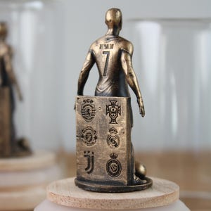May include: A bronze-colored figurine of a soccer player, jersey number 7, with the name "Ronaldo" on the back. The figurine stands on a wooden base and holds a panel with team logos. The figurine is approximately 4 inches tall.