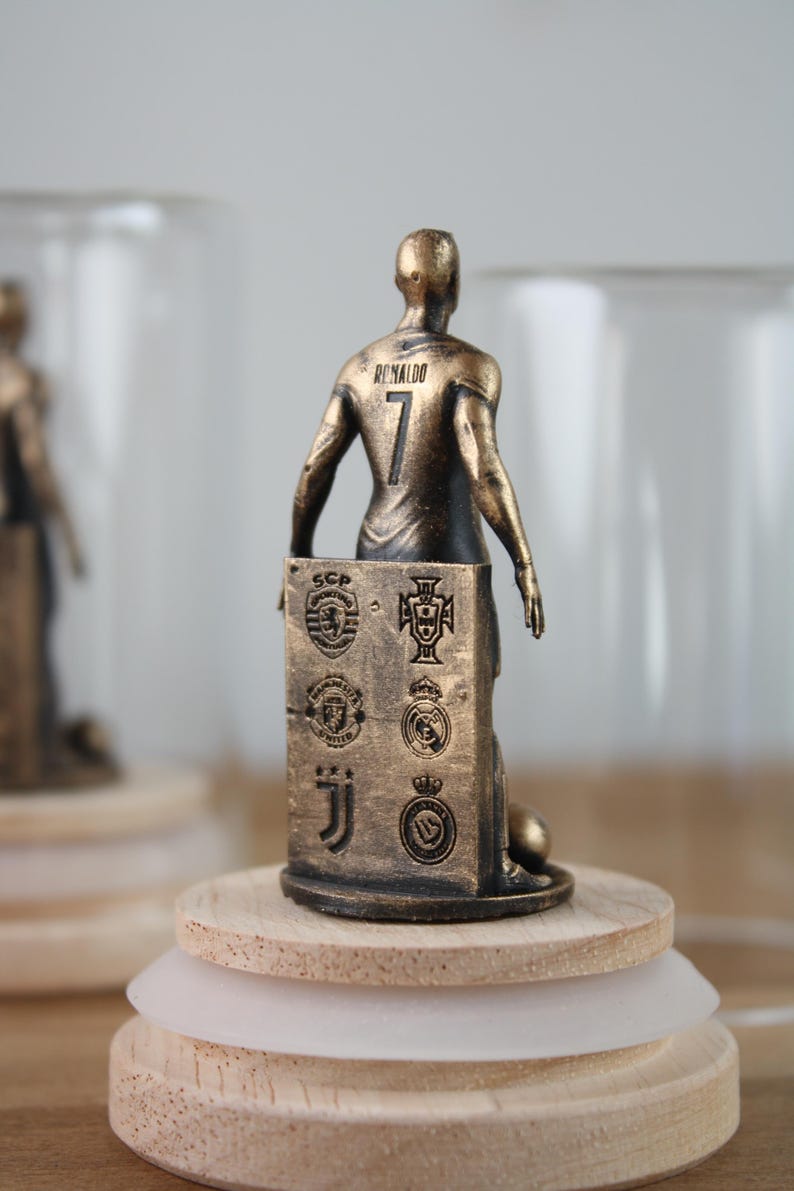 May include: A bronze-colored figurine of a soccer player, jersey number 7, with the name "Ronaldo" on the back. The figurine stands on a wooden base and holds a panel with team logos. The figurine is approximately 4 inches tall.