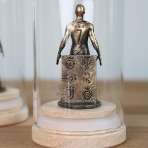 May include: A bronze-colored figurine of a soccer player, wearing the number 7 and the name "Ronaldo" on the back, displayed under a glass cloche. The figurine is mounted on a wooden base, with a block featuring team logos.
