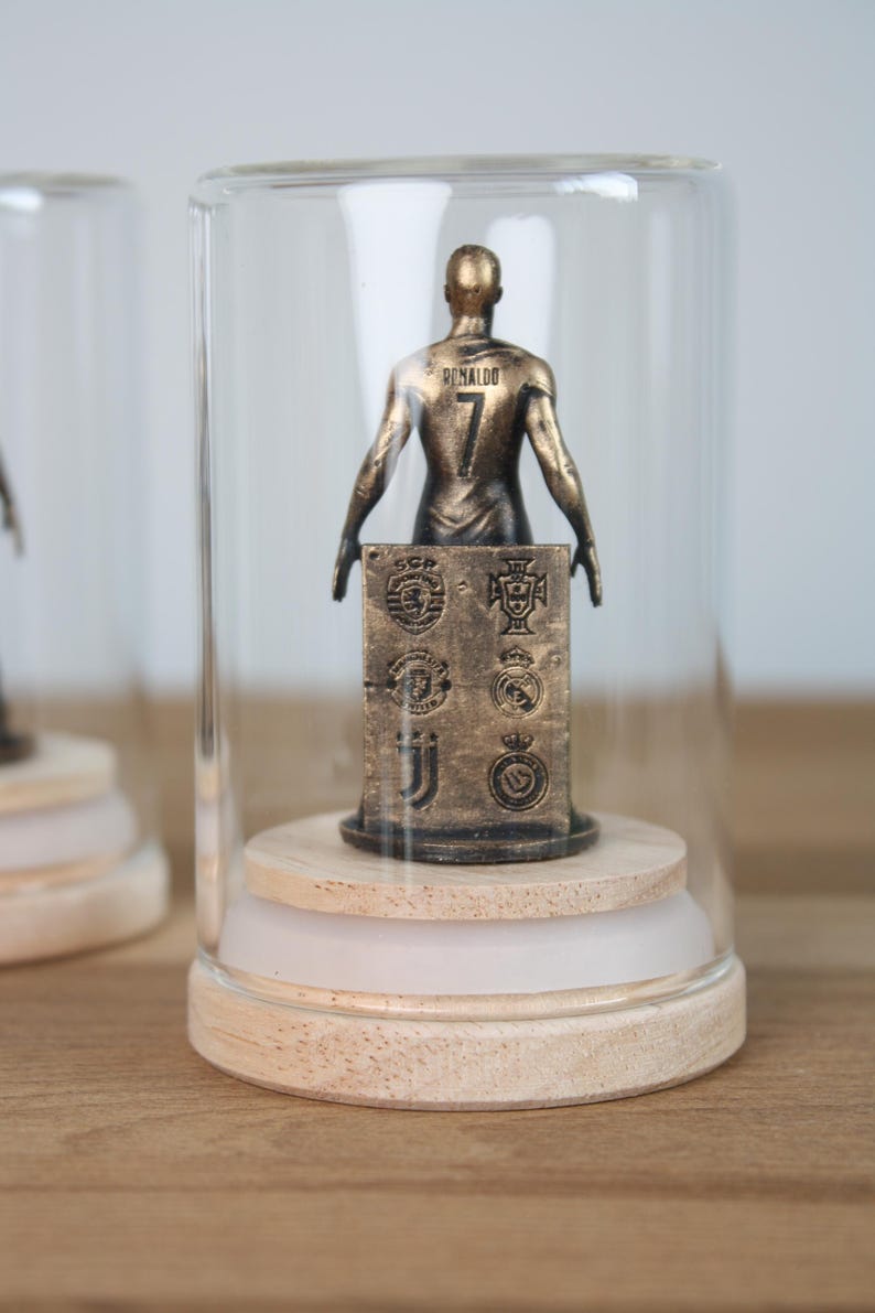 May include: A bronze-colored figurine of a soccer player, wearing the number 7 and the name "Ronaldo" on the back, displayed under a glass cloche. The figurine is mounted on a wooden base, with a block featuring team logos.
