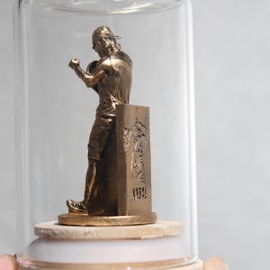 May include: A bronze-colored statue of a boxer inside a glass cloche. The statue is mounted on a wooden base with a white ring. The boxer is in a fighting stance, with one arm raised. The base has an inscription.