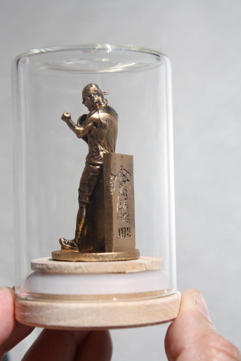 May include: A bronze-colored statue of a boxer inside a glass cloche. The statue is mounted on a wooden base with a white ring. The boxer is in a fighting stance, with one arm raised. The base has an inscription.