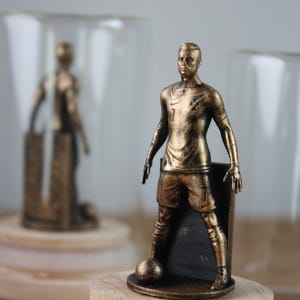 May include: A bronze-colored figurine of a soccer player in a uniform with the number 7. The figure is posed with a soccer ball at its feet. The figurine is mounted on a wooden base with a clear glass dome in the background.