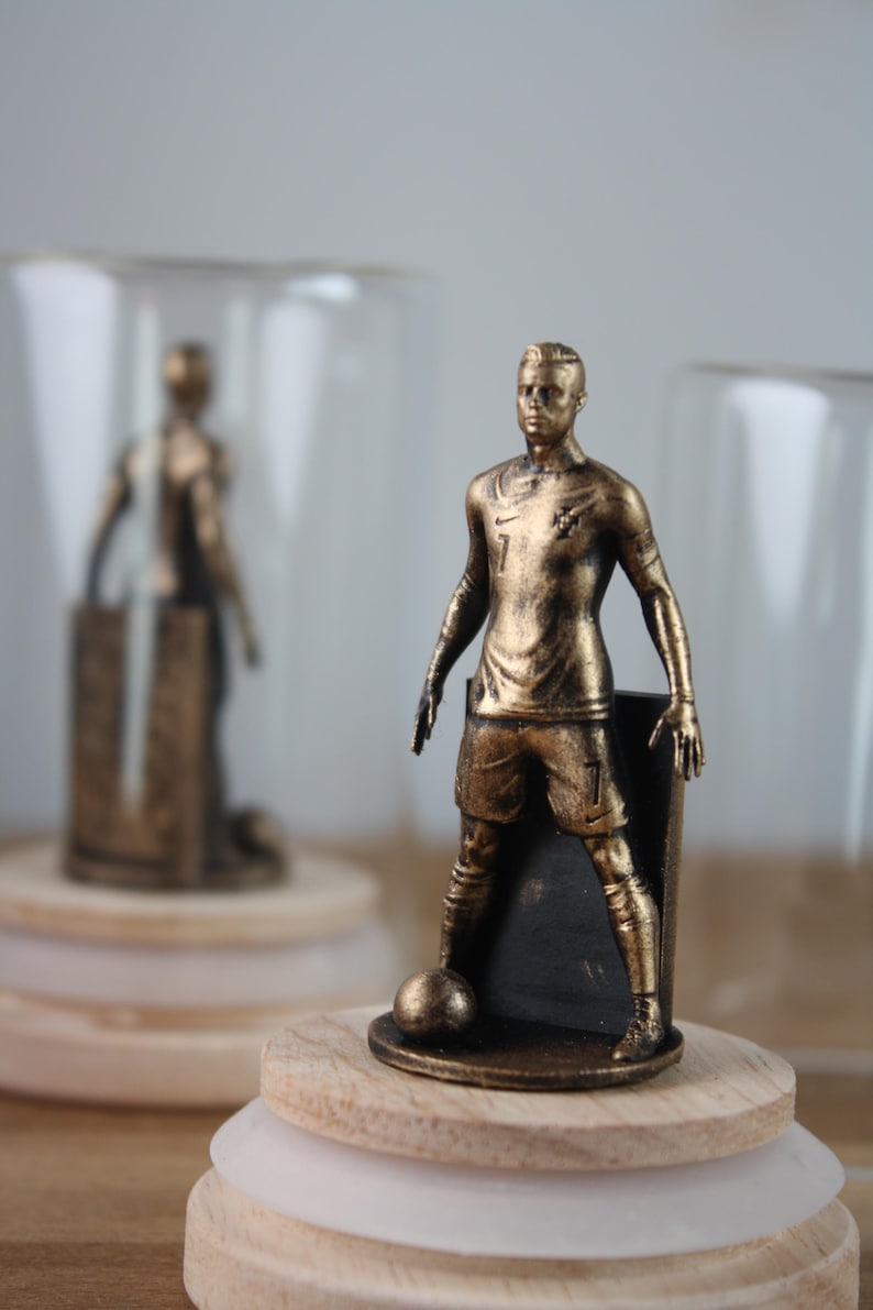 May include: A bronze-colored figurine of a soccer player in a uniform with the number 7. The figure is posed with a soccer ball at its feet. The figurine is mounted on a wooden base with a clear glass dome in the background.