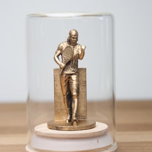 May include: A gold-colored tennis player figurine inside a glass cloche. The figurine is standing with a tennis racket and is mounted on a wooden base. The cloche is on a wooden surface.