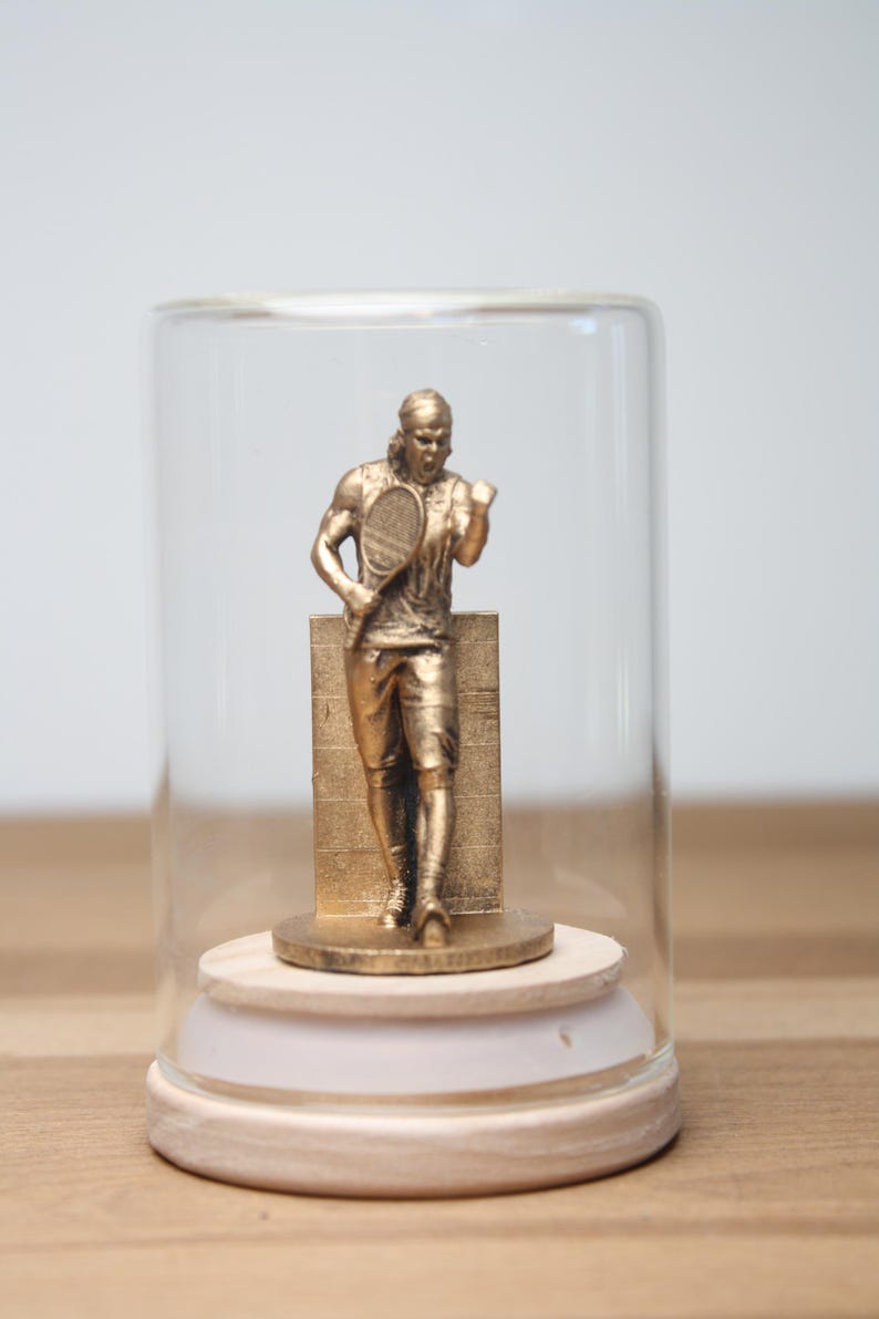 May include: A gold-colored tennis player figurine inside a glass cloche. The figurine is standing with a tennis racket and is mounted on a wooden base. The cloche is on a wooden surface.