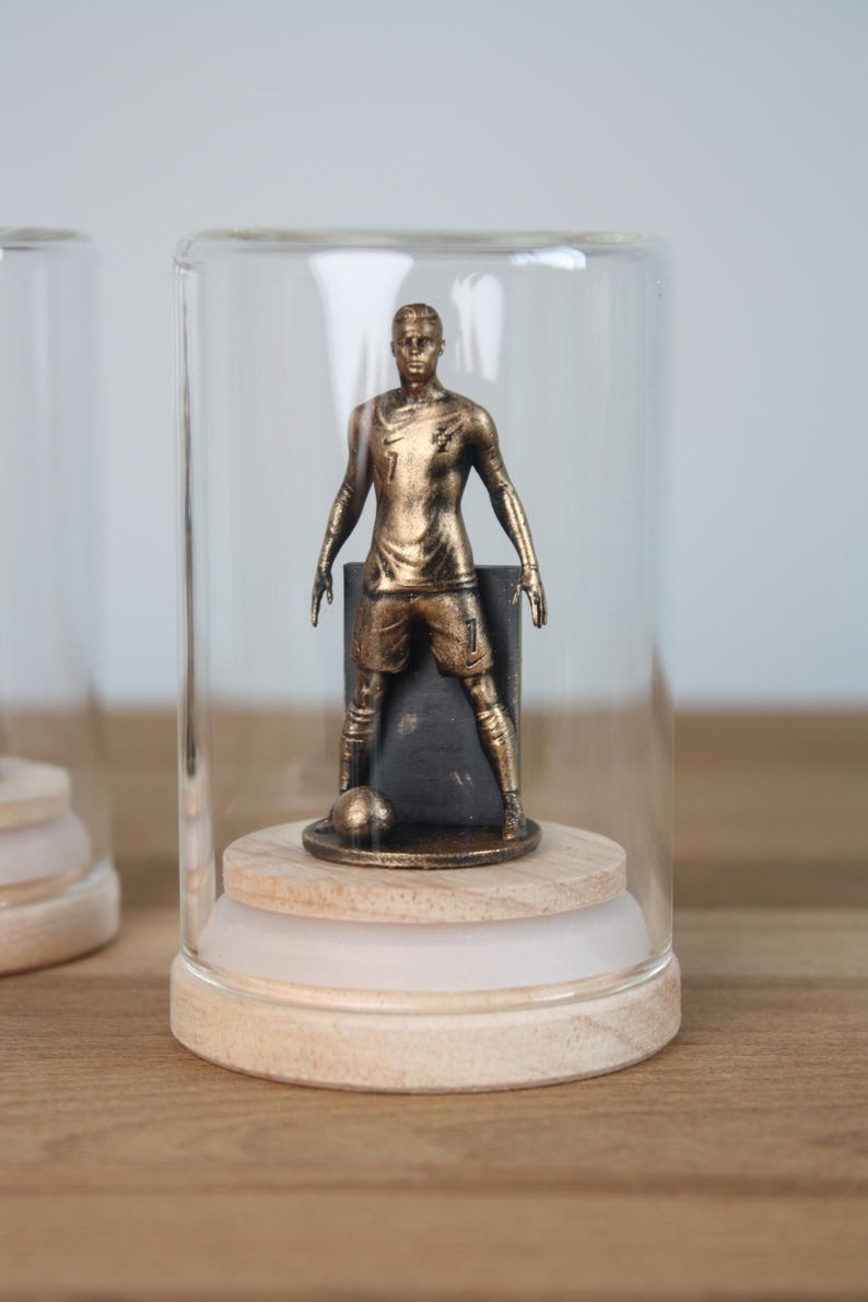 May include: A gold-colored soccer player figurine inside a glass dome. The figurine is on a wooden base with a soccer ball at its feet. The dome is clear, and the background is neutral.