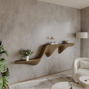 May include: A modern, wooden wall shelf with a unique, organic, wave-like design. The shelf is made of light brown wood and is mounted on a light gray wall. The shelf has a small, white vase with green plants on it.