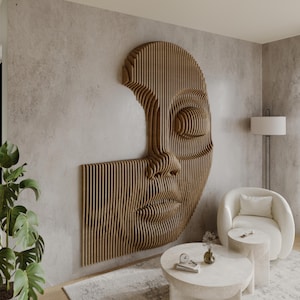 Puede incluir: Una escultura de pared de madera de un rostro, con un acabado marrón claro. La escultura está hecha de finas tiras de madera que están dispuestas en un patrón geométrico. La escultura está montada en una pared gris claro.