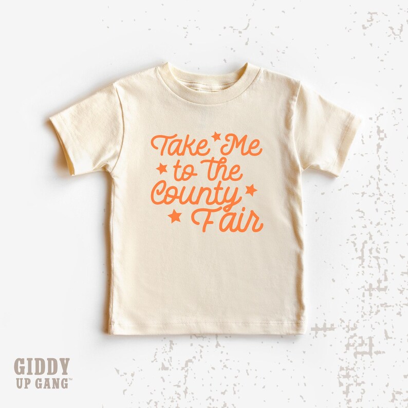 May include: A cream colored short-sleeved t-shirt with the text "Take Me to the County Fair" in orange.