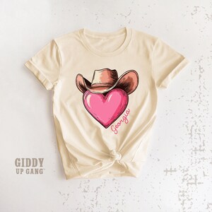 May include: Cream-colored t-shirt featuring a graphic of a cowboy hat over a pink heart with the word "Georgia" in cursive. The shirt has short sleeves and a knotted hem. The text "GIDDY UP GANG" is printed on the lower left.