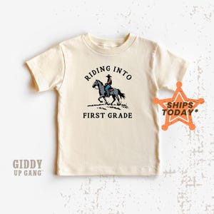 May include: A beige short-sleeved t-shirt with the text "RIDING INTO FIRST GRADE" and a cartoon image of a cowboy riding a horse.