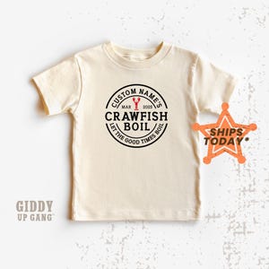 May include: Cream-colored t-shirt with a circular graphic that reads "CUSTOM NAME'S CRAWFISH BOIL" with a crawfish illustration and "LET THE GOOD TIMES BOIL." The shirt also has a star-shaped graphic that says "SHIPS TODAY."