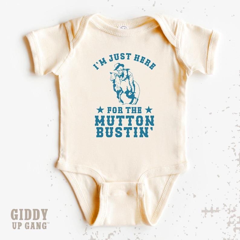 May include: A cream-colored baby onesie with a blue graphic of a cowboy riding a sheep. The text reads "I'm just here for the mutton bustin'" with stars on either side of the text.
