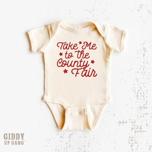 May include: A cream-colored baby onesie with red text that reads "Take Me to the County Fair" with red stars.