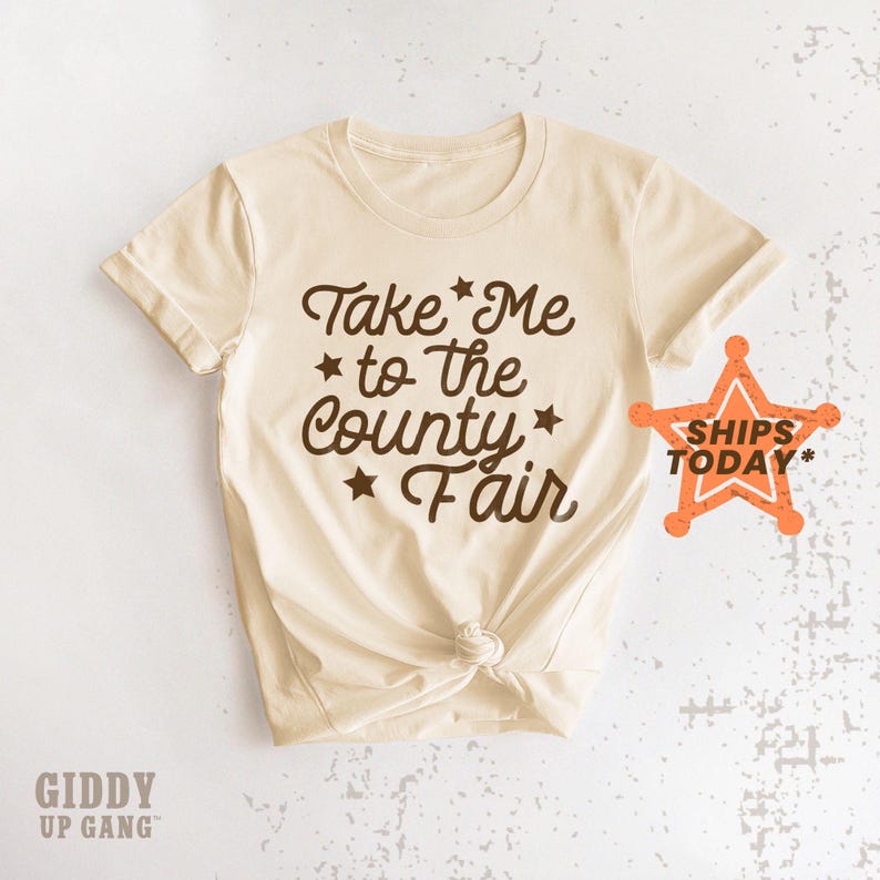 May include: A beige t-shirt with brown text that reads "Take Me to the County Fair" with stars around the text. The shirt is knotted in the front and has a distressed look.