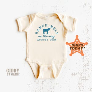 May include: A cream-colored baby onesie with a blue graphic that says "Ranch Help on the way August 2024" and a silhouette of a cow. The onesie has short sleeves and a snap closure.
