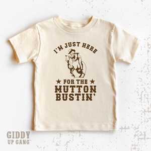 May include: A beige t-shirt with a brown graphic of a cowboy riding a sheep. The text on the shirt reads "I'm just here for the mutton bustin'".