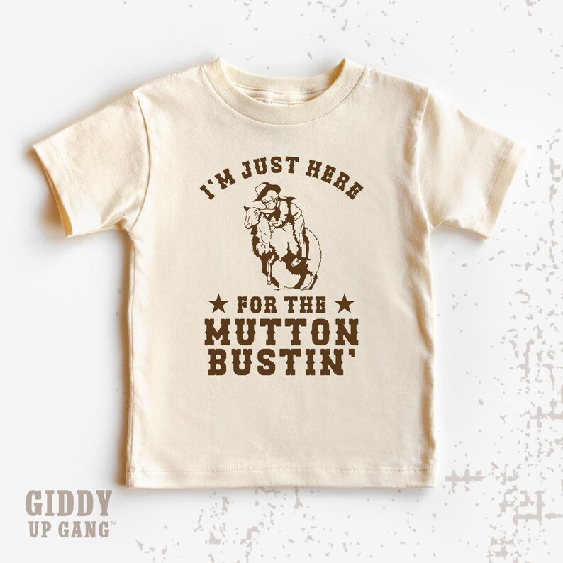 May include: A beige t-shirt with a brown graphic of a cowboy riding a sheep. The text on the shirt reads "I'm just here for the mutton bustin'".