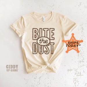 May include: Cream-colored t-shirt with the phrase "Bite the Dust" in a brown rope-like font. The shirt has a crew neck and short sleeves. A star-shaped graphic with the words "Ships Today" is on the right side. The brand "Giddy Up Gang" is printed on the bottom left.