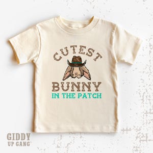 May include: Cream-colored t-shirt featuring a graphic of a bunny wearing a cowboy hat. The text above the bunny reads "CUTEST" and below it, "BUNNY IN THE PATCH" in brown and teal lettering. The brand name "GIDDY UP GANG" is printed at the bottom.