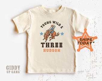 Könnte beinhalten: Ein beiges T-Shirt mit einer Grafik im Vintage-Stil, die einen Cowboy auf einem buckelnden Pferd zeigt. Der Text "Young Wild & Three Hudson" ist unter der Grafik gedruckt.