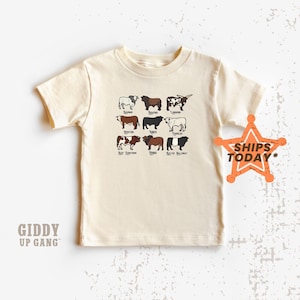 May include: A cream-colored t-shirt with a graphic of different breeds of cattle. The breeds are labeled with their names: Belted Galloway, Red Devon, Hereford, Angus, Charolais, Longhorn, Shorthorn, and Brahman. The text "Giddy Up Gang" is at the bottom of the shirt.