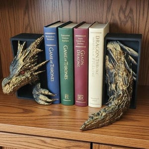 May include: Dragon-themed bookends with a gold and black finish, holding a collection of books. The books include titles like "Game of Thrones" and "Dragon Throne". The bookends are on a wooden shelf.