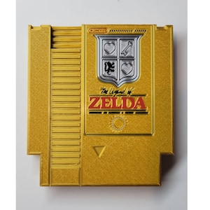 May include: Gold Nintendo Entertainment System (NES) game cartridge for The Legend of Zelda. The cartridge features a silver shield with a heart, key, lion, and heart design. The title "The Legend of Zelda" is printed in red and black.