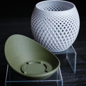 May include: A white, lattice-patterned decorative vase and an olive green, oval-shaped planter. The vase has a spherical shape with a diamond-shaped pattern. The planter has a textured surface and a circular base.