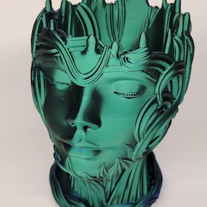 May include: A green ceramic planter shaped like a human face with closed eyes and a detailed, textured surface.