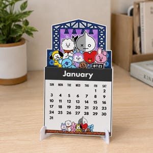 May include: A desktop calendar for January featuring BT21 characters on a stage graphic at the top. The calendar has a white background with black text for the days of the week and dates. Small colorful character illustrations are at the bottom.