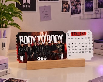 Könnte beinhalten: Ein rechteckiger Tischkalender im Ticket-Design mit einem Foto von sieben Personen in schwarzer Kleidung und dem Text BODY TO BODY. Der Kalender zeigt den Monat Januar 2027 und steht auf einem hellen Holzsockel.