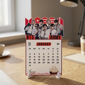 Può includere: Un calendario da scrivania 2027 con una fotografia di gruppo di sette persone in camicia bianca e cravatta nella parte superiore. La pagina del calendario mostra il mese di gennaio con il testo BTS ARMY, la parola Swim e un veliero illustrato.