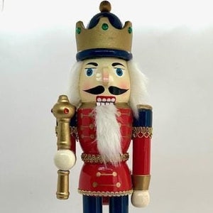 May include: A wooden nutcracker figurine wearing a red and blue uniform, a gold crown, and a white beard. The nutcracker is holding a gold sceptre and stands on a gold base.