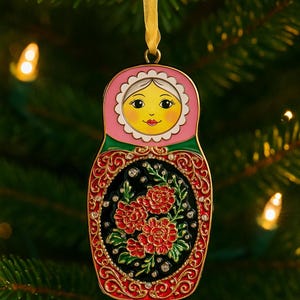 May include: A decorative Christmas ornament shaped like a Matryoshka doll. The ornament features a pink head with a smiling face, a green and red body, and a black center with red floral details. It hangs from a gold ribbon.