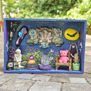 May include: A blue shadow box with a variety of miniature gothic-themed items. Includes a pink teddy bear on a gold chair, a skeleton, a tombstone with "RIP", and a yellow moon with a bat. Other items include a bottle with green beads and a purple crystal.