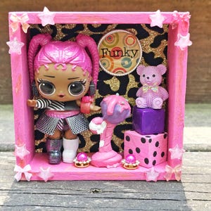 May include: A pink shadow box featuring a doll with pink hair, a teddy bear, and a lollipop. The background has a gold and black leopard print. The box is decorated with star-shaped embellishments and the word "Funky".