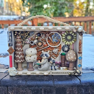 May include: A rectangular wooden shadow box frame featuring a decorative collage of two raccoon figurines, wooden mushroom shapes, and various vintage jewelry pieces including floral brooches, beads, and metal charms, finished with a jute twine handle.