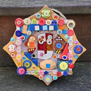May include: A mixed media art piece featuring a geometric design with a central face-like composition. The artwork incorporates various colorful buttons, wooden shapes, and decorative elements, creating a textured and vibrant aesthetic. The piece is mounted on a wooden star-shaped frame.