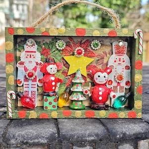 May include: A Christmas-themed shadow box with a green and red polka-dot frame. Inside are miniature nutcrackers, a Christmas tree, and a star, with decorative jewels and candy canes. A jute rope is attached for hanging.