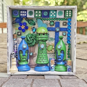 May include: A decorative shadow box with a white distressed frame. The box contains various objects, including dice, chess pieces, and decorative bottles. The chess pieces are painted blue and green. Embellishments include blue and green gems and beads.