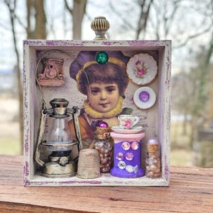 May include: A small wooden box with a purple and white painted finish. The box contains a variety of miniature items, including a lantern, a thimble, a spool of thread, and a small teacup and saucer. The box is decorated with a vintage image of a young girl.