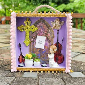 May include: A decorative religious shadow box with a purple interior, featuring a glittery cross, a stained-glass window, and a miniature figure of a praying girl. The box includes a small violin, a cello, and the text "Holy Bible".