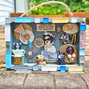May include: A blue wooden box with a glass front, decorated with a variety of colorful buttons, beads, and other trinkets. The box has a sign that reads "Travel the world to find the beautiful".