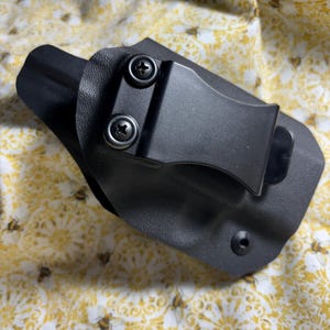 May include: A black, molded polymer holster with a metal belt clip and two screws. The holster is designed to be worn on the inside of the waistband. The image is taken from an overhead perspective.