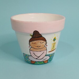 May include: A white ceramic flower pot with a pink rim, hand-painted with a cartoon illustration of a person meditating with a candle and the word "MEAN" on the side.